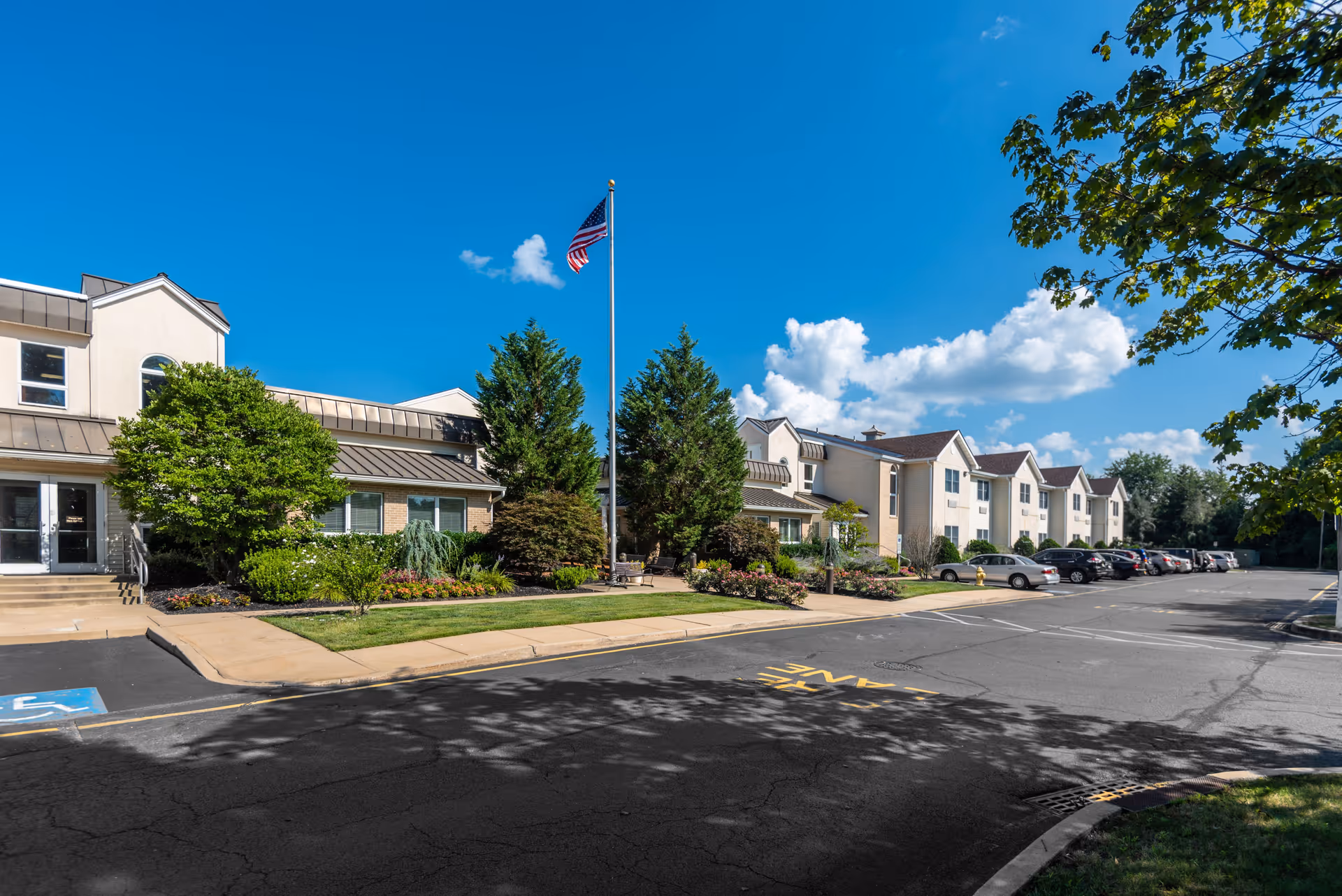 Exterior view of a senior living facility building with a parking lot, landscaped greenery, and an American flag on a flagpole under a clear blue sky with some clouds.