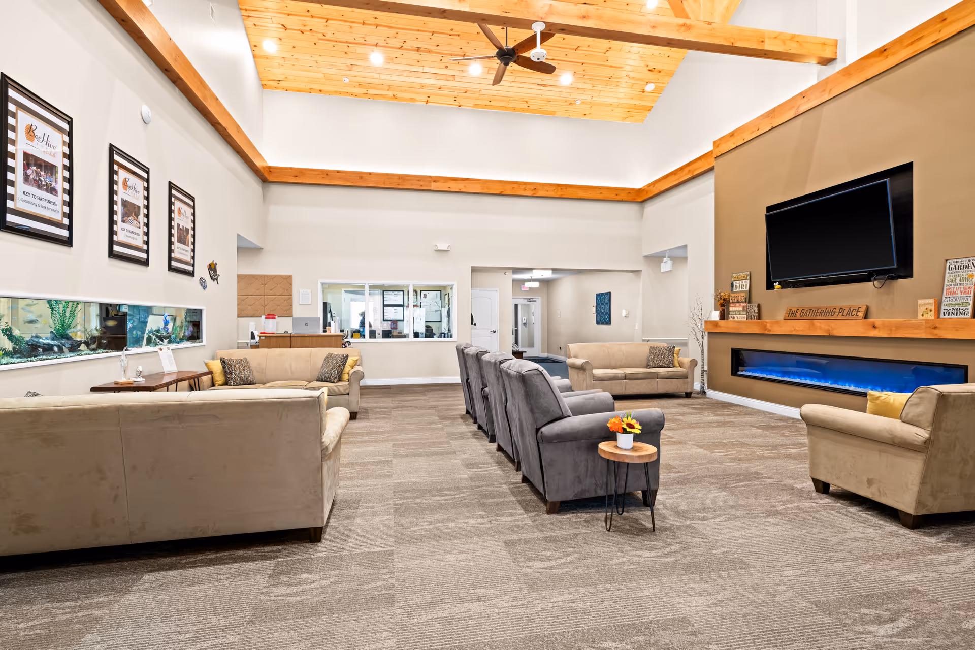 Spacious assisted living community lounge with sofas and armchairs facing a wall-mounted TV and long electric fireplace under a high wooden-beamed ceiling.