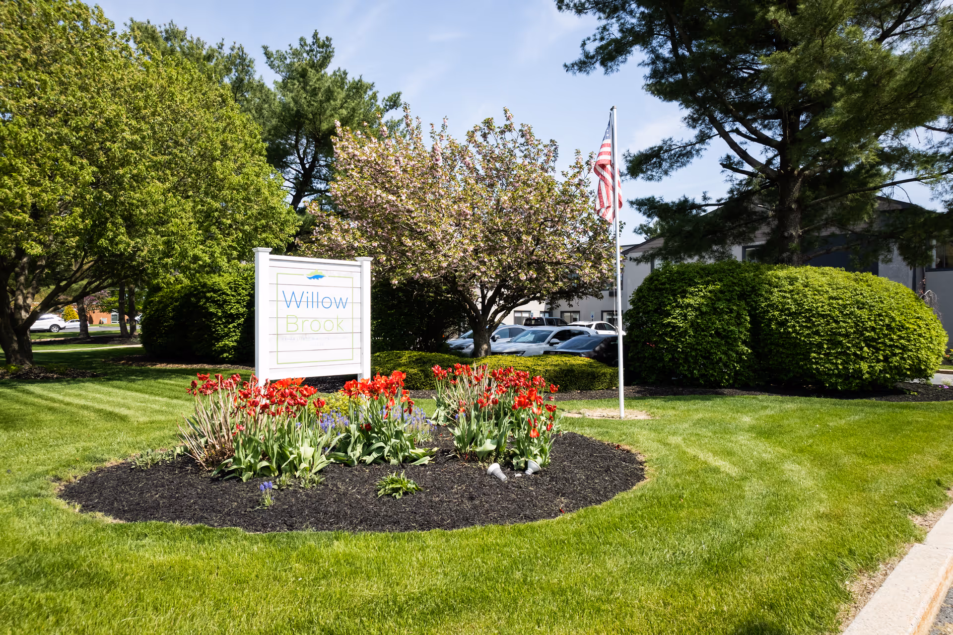 A landscaped outdoor area with a flower bed containing red tulips and other plants surrounding a white sign that reads 'Willow Brook'. Behind the flower bed is a flagpole with an American flag, green bushes, trees, and a parking lot with cars visible in the background under a clear blue sky.