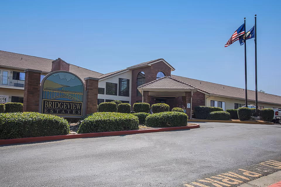Exterior view of Bridgeview Estates senior living facility with a large sign displaying the facility name, well-maintained bushes, a paved driveway, and two flagpoles flying the American and state flags under a clear blue sky.