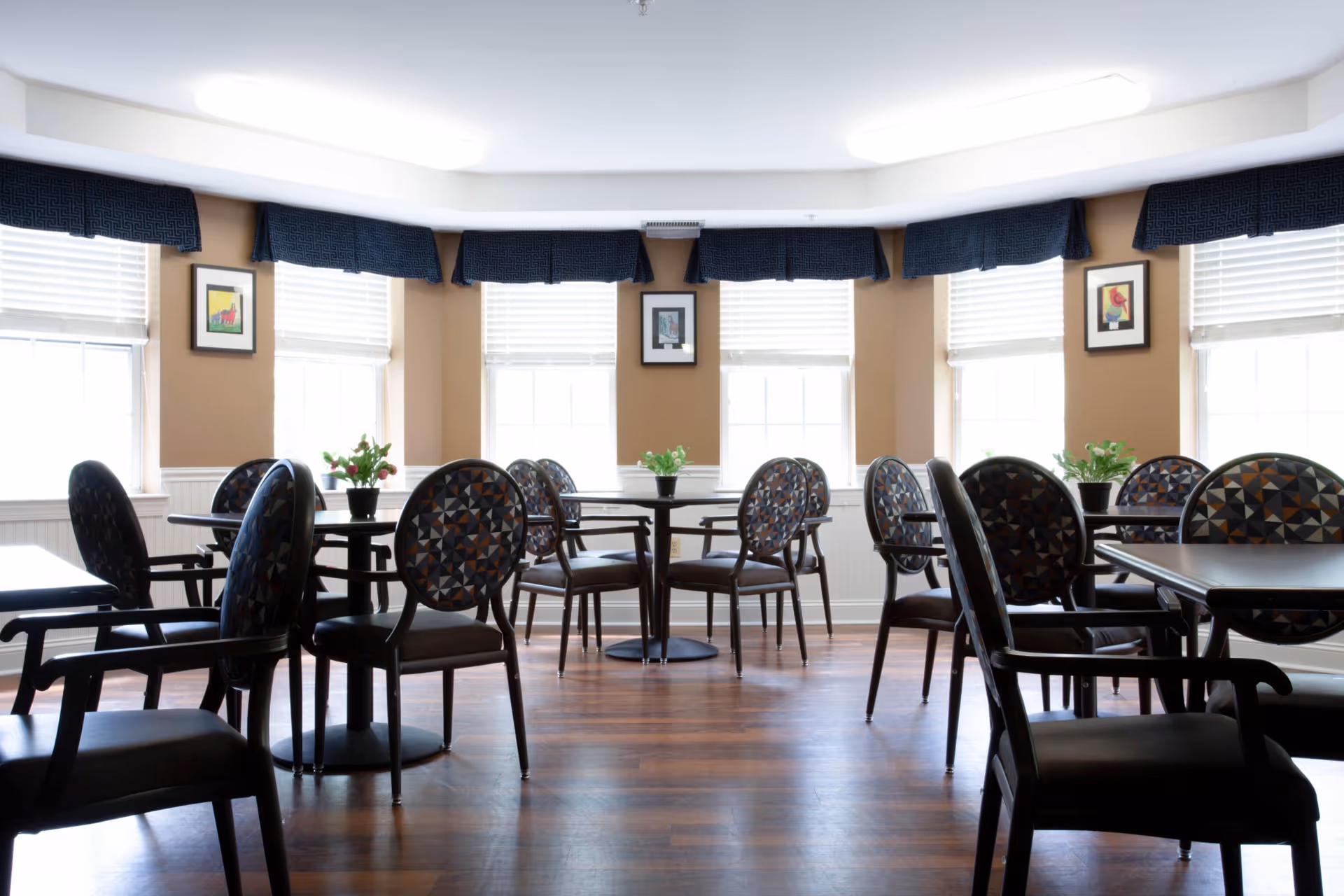 Well-lit dining room with round tables and patterned chairs arranged on hardwood floors beneath large windows.