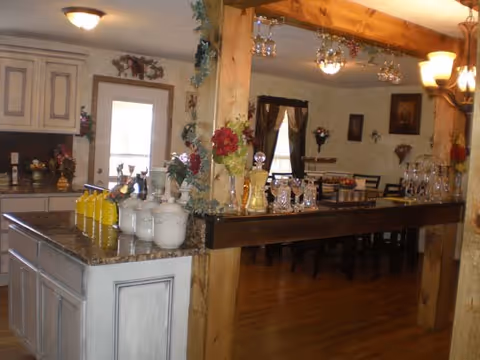 Open kitchen with a central island topped with jars and decorative flowers, wooden beams, and a dining area with a long table and chairs in the background.
