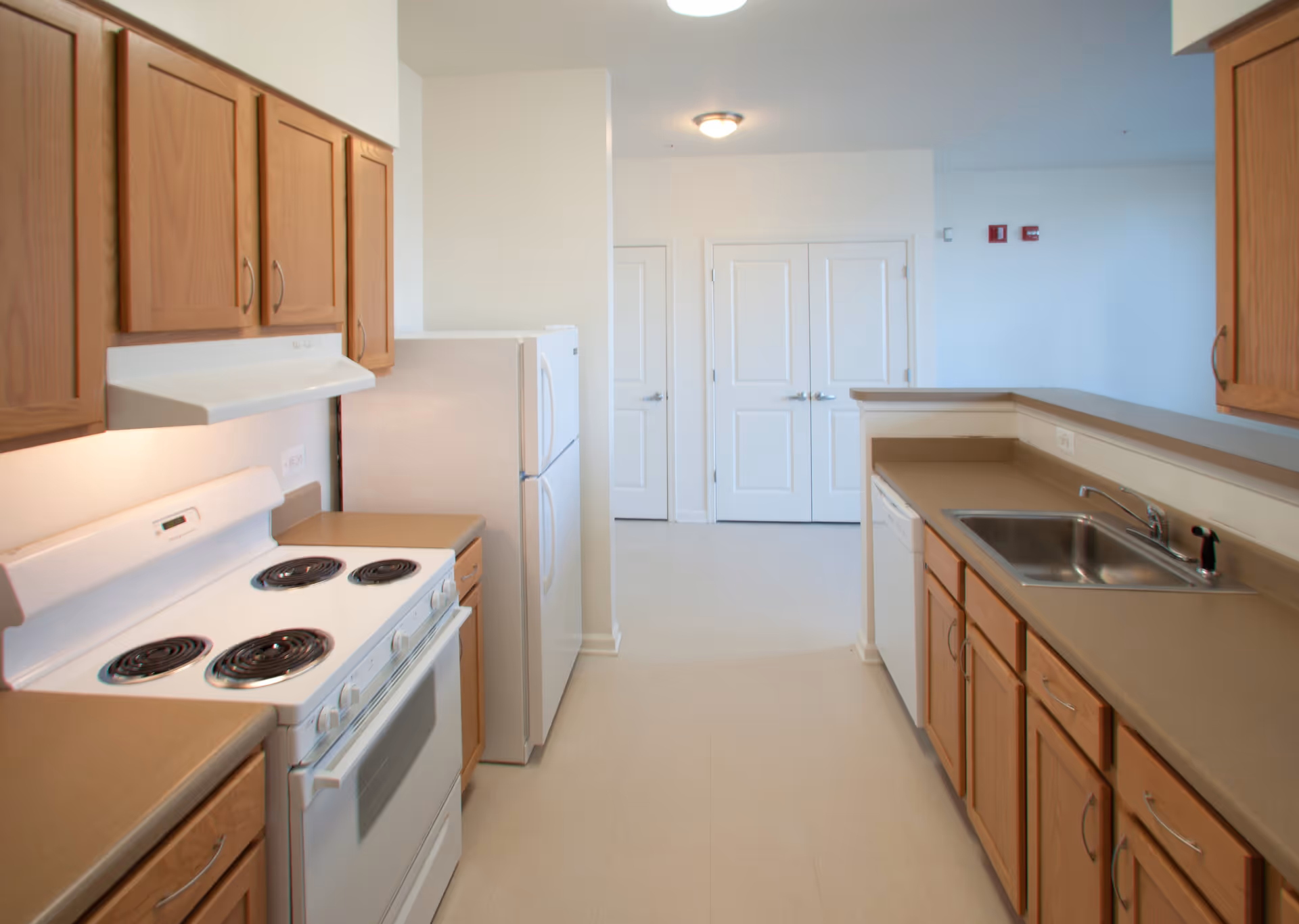 A clean, modern kitchen with wooden cabinets, a white electric stove with four coil burners, a white refrigerator, a dishwasher, and a stainless steel sink. The kitchen has beige countertops and light-colored flooring, with white walls and two closed white doors in the background.