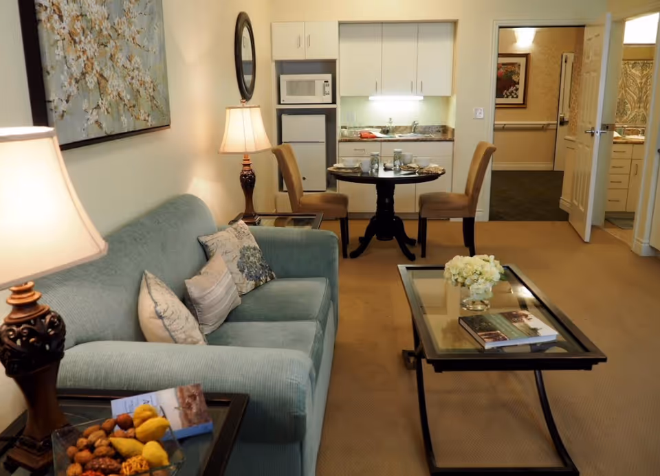 A cozy senior living room with a light blue sofa adorned with decorative pillows, two table lamps on side tables, a glass coffee table with a flower vase and a book, and a small dining area with a round table set for two. In the background, there is a kitchenette with a microwave, mini fridge, and cabinets, and an open door leading to another room.