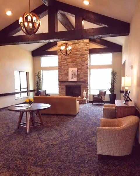 A spacious living room with high vaulted ceilings featuring dark wooden beams and two spherical chandeliers. The room has a stone fireplace centered on the far wall with a framed picture above it. There are two tall plants flanking the fireplace and two armchairs placed near it. In the middle of the room is a beige sofa facing the fireplace. A round wooden table with a small flower arrangement is positioned on the left side, and two beige armchairs with a wooden side table and lamp are on the right side. Large windows allow natural light to brighten the room.