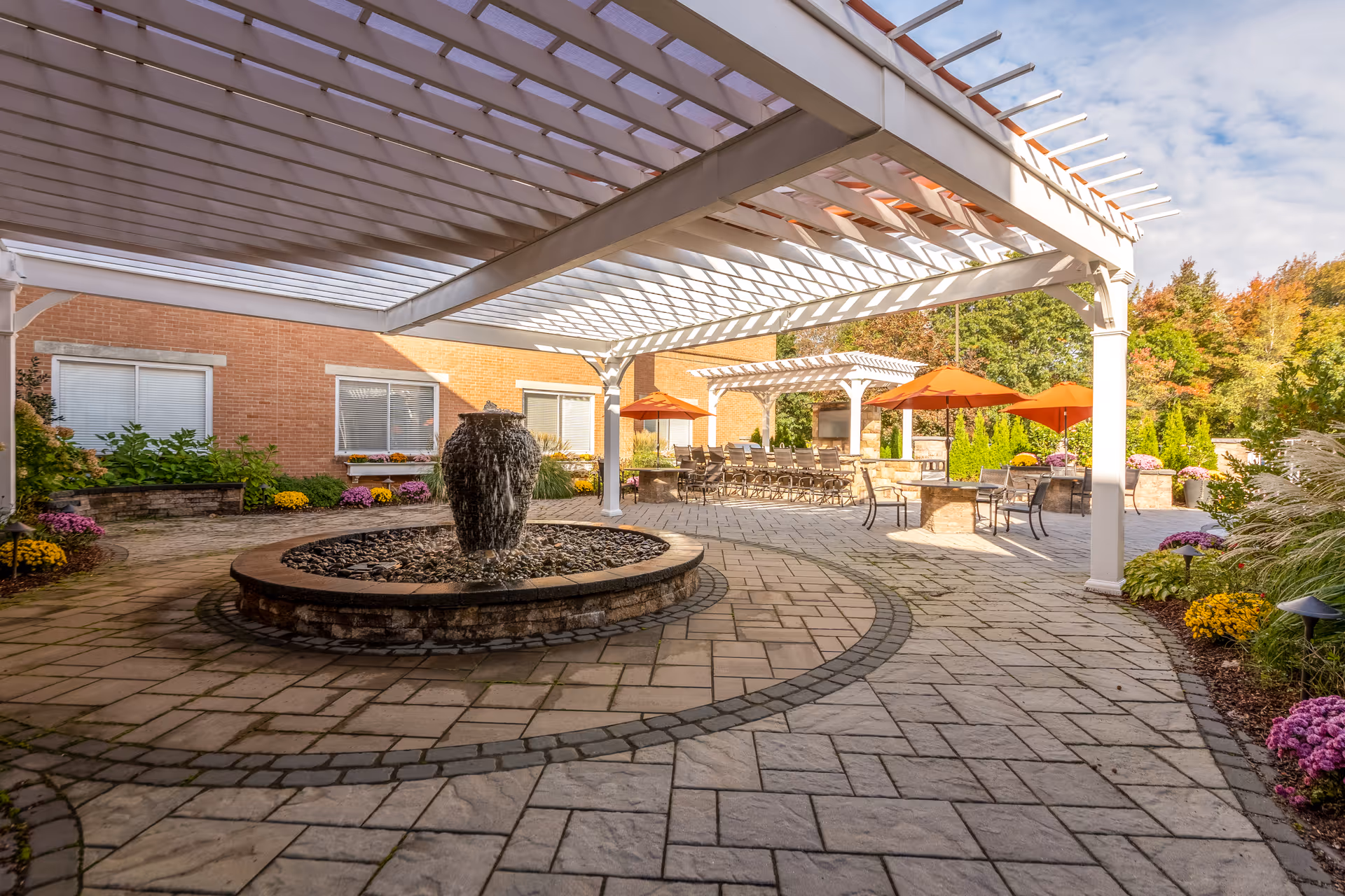 Outdoor patio area at CareOne at Weymouth featuring a stone fountain in the center, surrounded by a paved stone floor. There are white pergolas providing partial shade, several tables with orange umbrellas, chairs, and landscaped flower beds with colorful flowers and greenery. The background includes a brick building and trees with autumn foliage.