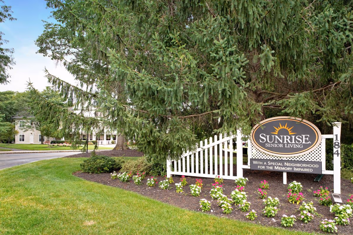 Outdoor view of the entrance sign for Sunrise Senior Living, surrounded by green grass, flower beds, and large evergreen trees, with a building visible in the background.