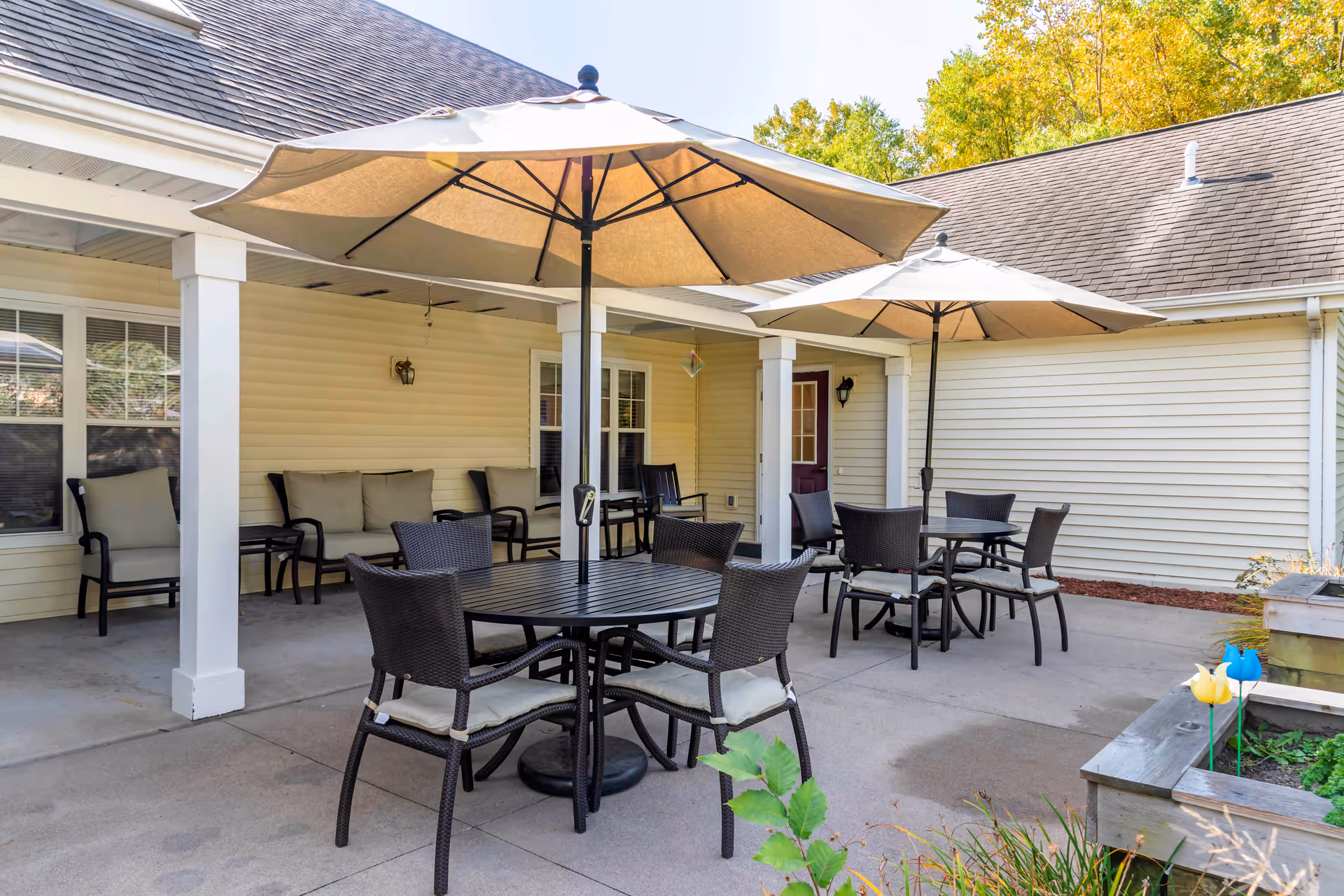 Outdoor patio area with two round tables, each surrounded by four wicker chairs with cushions, and large beige umbrellas providing shade. There is a covered porch with cushioned seating along the wall of a light yellow building with white trim. Some greenery and a raised garden bed with decorative pinwheels are visible in the foreground.