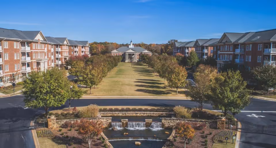 A wide view of the Cascades Verdae senior living facility showing two large brick residential buildings on either side of a long, grassy courtyard lined with trees. In the foreground, there is a landscaped area with a small waterfall feature and a stone wall. The sky is clear and blue.