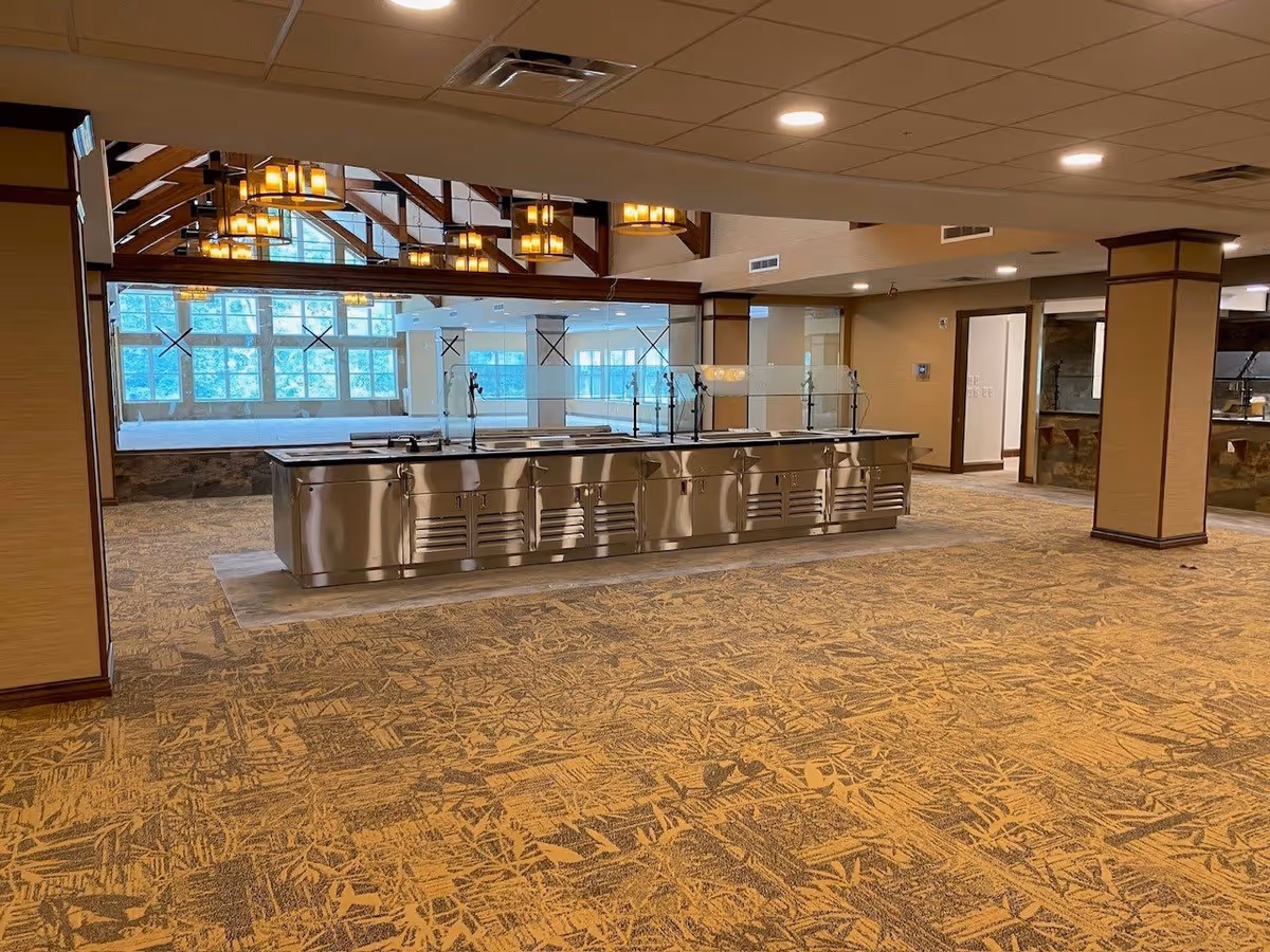 Spacious dining room with a central stainless-steel buffet counter, patterned carpet, exposed wood beams and pendant lights.