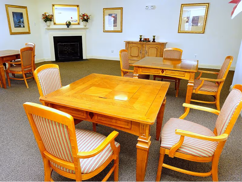 A well-lit room with wooden tables and striped cushioned chairs arranged for seating. The room features a fireplace with a white mantel, decorated with flower arrangements and framed pictures on the walls.