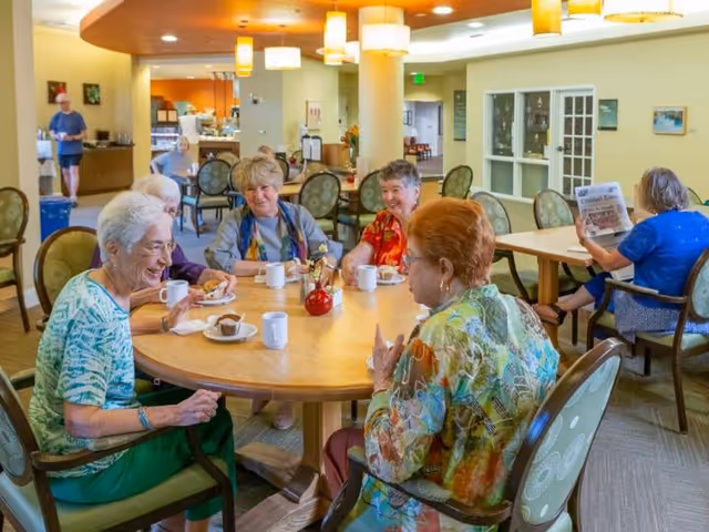 A group of elderly women sitting around a round wooden table in a well-lit dining area, enjoying coffee and pastries. One woman is reading a newspaper in the background. The room has warm lighting with hanging lamps and several chairs and tables.