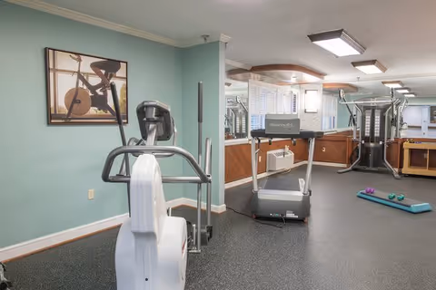 A fitness room with exercise equipment including a treadmill, elliptical machine, and multi-station gym. The room has light green walls, a framed picture of a person on a stationary bike, large mirrors, and a window with white blinds. There are also small weights and an exercise step on the floor.
