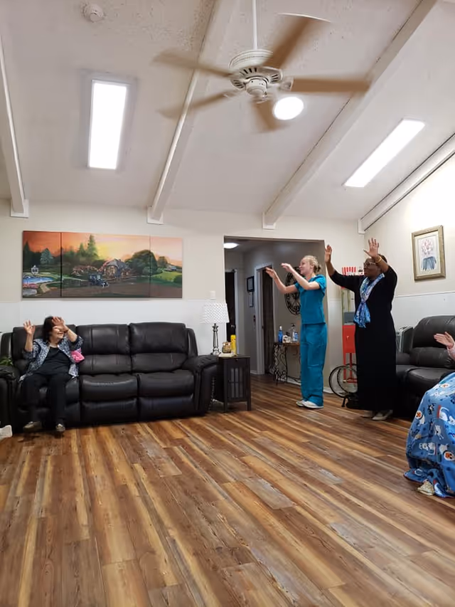 A group of people in a living room with wooden flooring and a ceiling fan. Two women are standing and raising their hands, while others are seated on dark leather couches. The room has a painting on the wall, a side table with a lamp, and bright overhead lights.