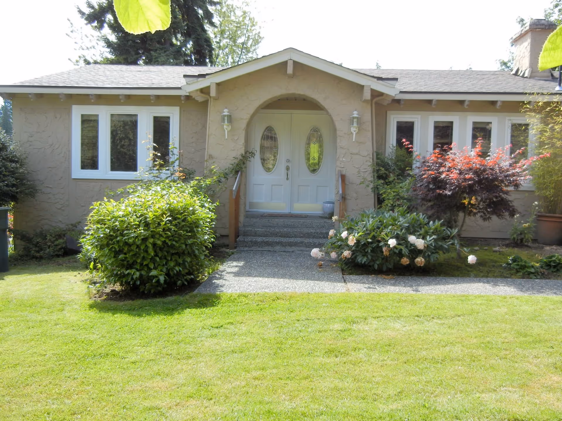 Front exterior view of a single-story house with beige stucco walls, white double front doors with oval glass panels, and a small porch with steps. The house is surrounded by green bushes, flowering plants, and a well-maintained lawn.