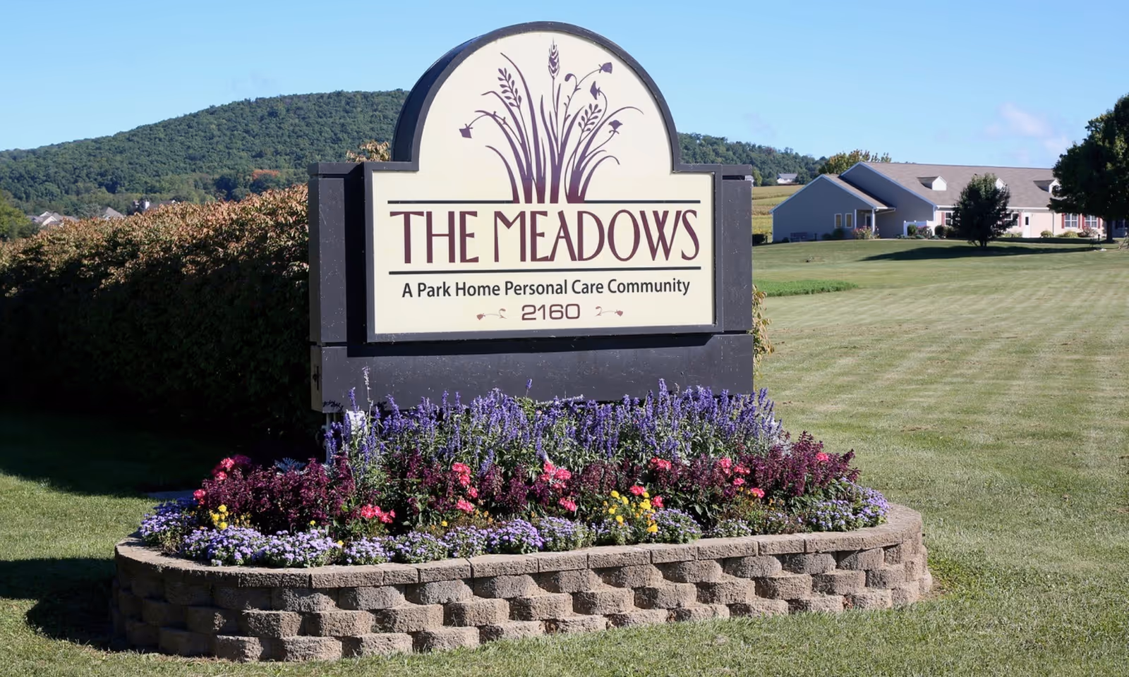 A large outdoor sign for The Meadows, A Park Home Personal Care Community, surrounded by a flower bed with purple, pink, and yellow flowers. The sign is set on a stone base with a grassy lawn and buildings in the background under a clear blue sky.