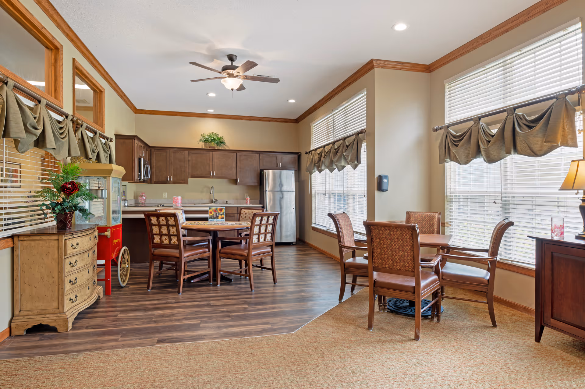 Bright communal dining area with round tables and chairs next to a small kitchen with cabinets and stainless steel refrigerator.