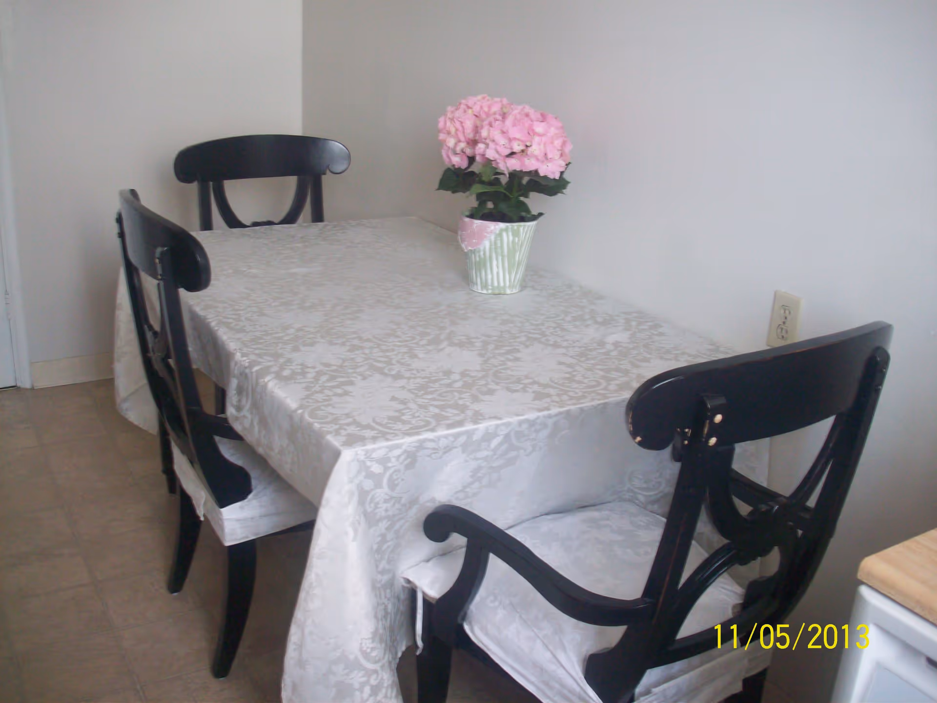 A dining area with a rectangular table covered by a white patterned tablecloth. There are four black wooden chairs with white cushions around the table. A small potted plant with pink flowers is placed in the center of the table. The room has plain white walls and a tiled floor.