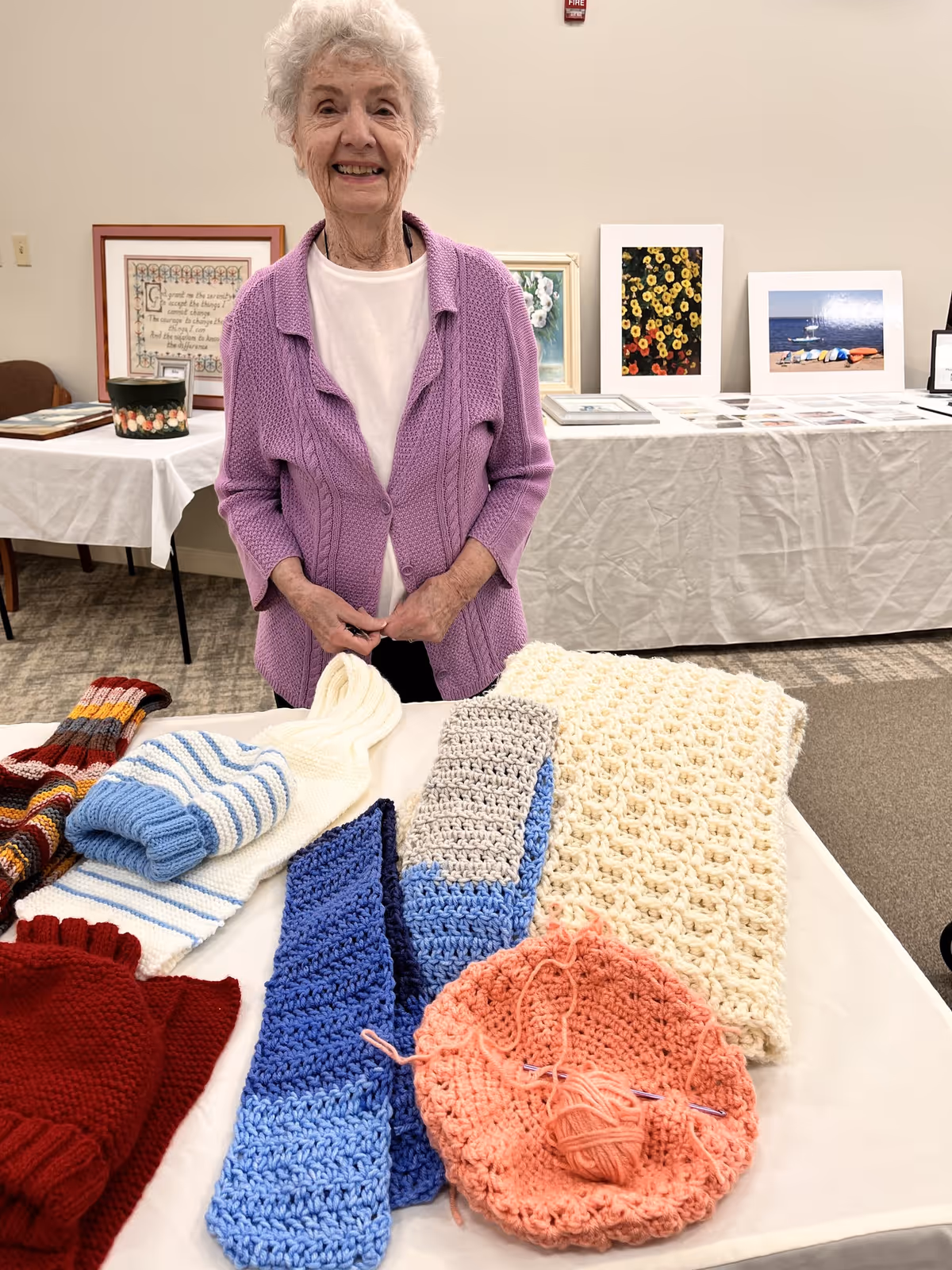 An elderly woman wearing a purple cardigan stands behind a table displaying various knitted and crocheted items including scarves, a hat, and a blanket. Behind her, there is a table covered with a white cloth displaying framed artwork and photographs.