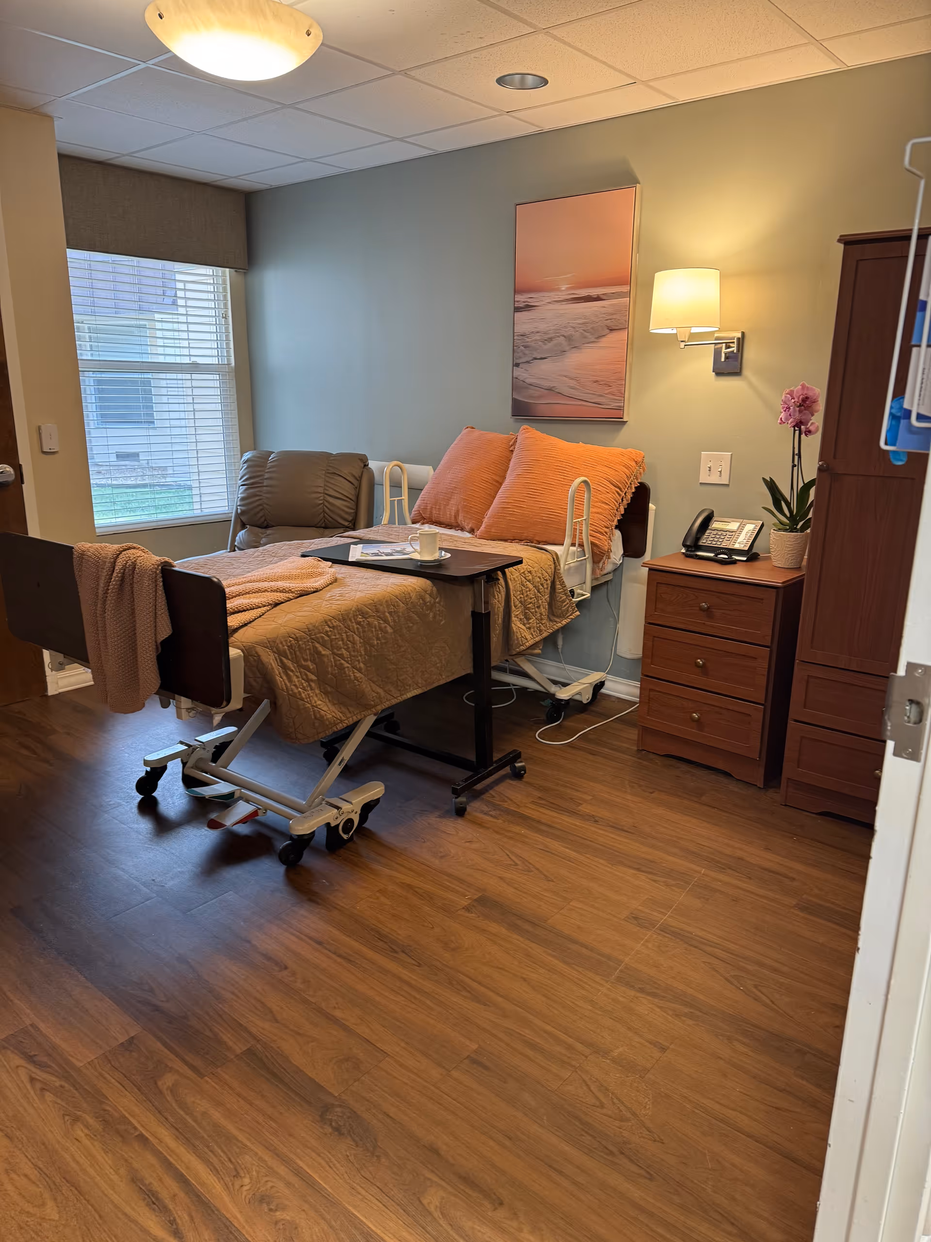 A senior living facility room featuring a bed, armchair, and bedside table.