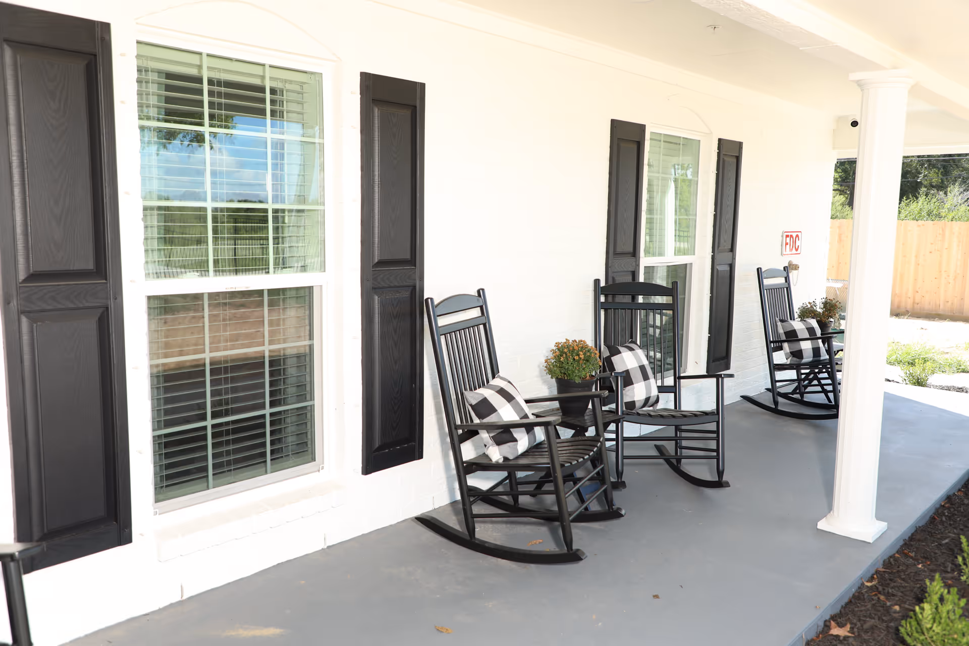 Covered front porch of a white building with black rocking chairs, checkered pillows, and potted plants.