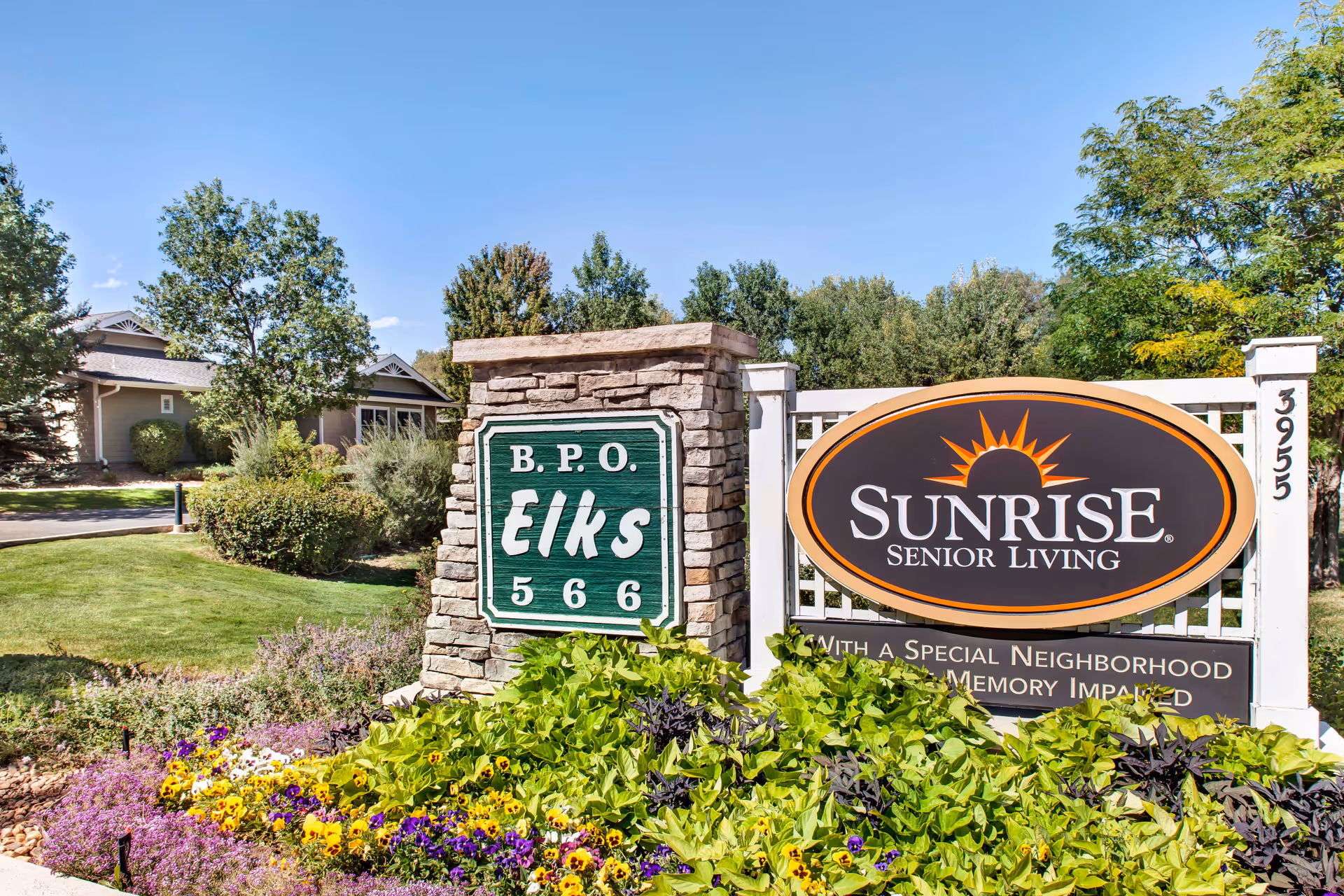 Outdoor view of the entrance sign for Sunrise Senior Living at B.P.O. Elks 566, surrounded by green plants, colorful flowers, and trees under a clear blue sky.