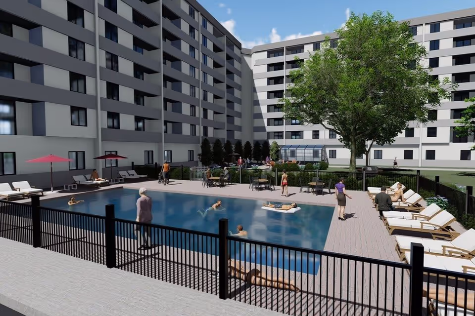 Outdoor swimming pool area at a senior living facility with people swimming, lounging on chairs, and walking around. The pool is surrounded by a black fence, lounge chairs with umbrellas, and a large tree in the background near a multi-story building.