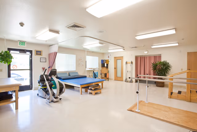 Bright rehabilitation room with parallel bars, an exercise bike, treatment tables, and therapy equipment.