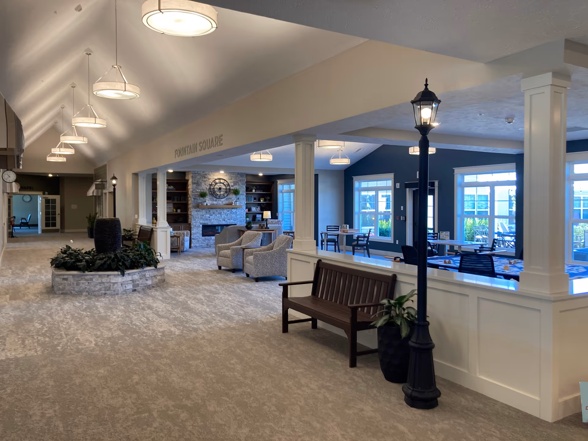 Spacious, well-lit senior living community interior with seating areas, a fireplace, dining tables, and decorative planters.
