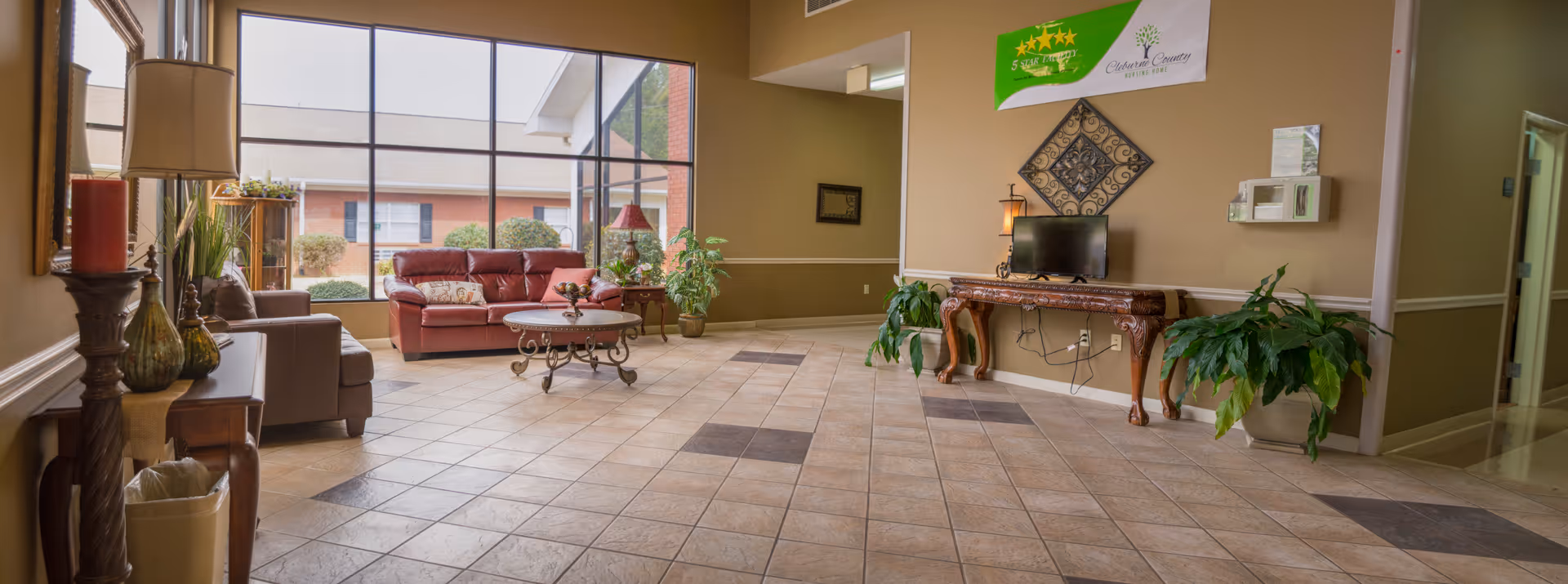 Spacious nursing-home lobby with tiled floor, seating area of sofas and a coffee table, large windows, plants, and a decorative console table against the wall.
