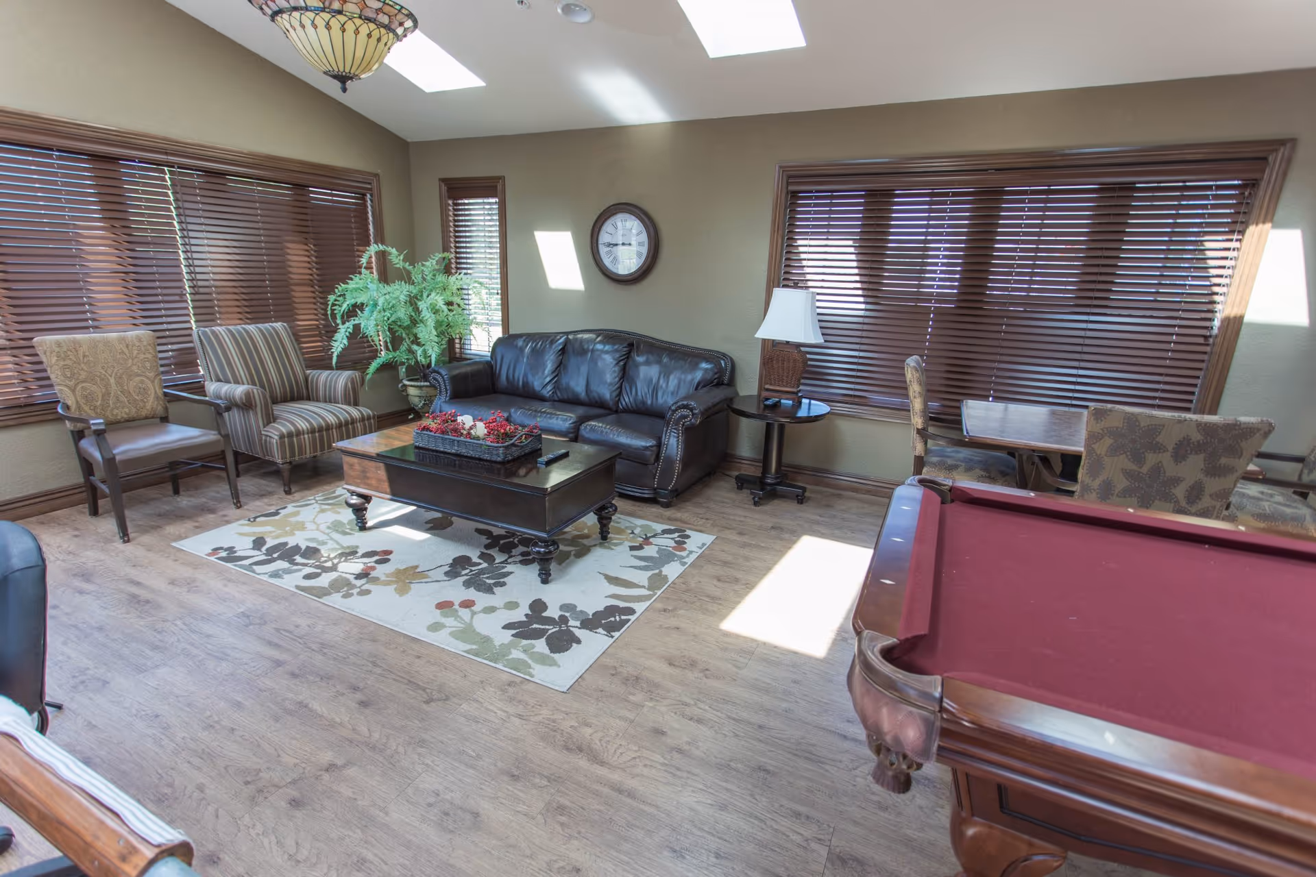 Sunlit lounge with a leather sofa, armchairs, coffee table on a patterned rug, and a pool table near large windows with wooden blinds.