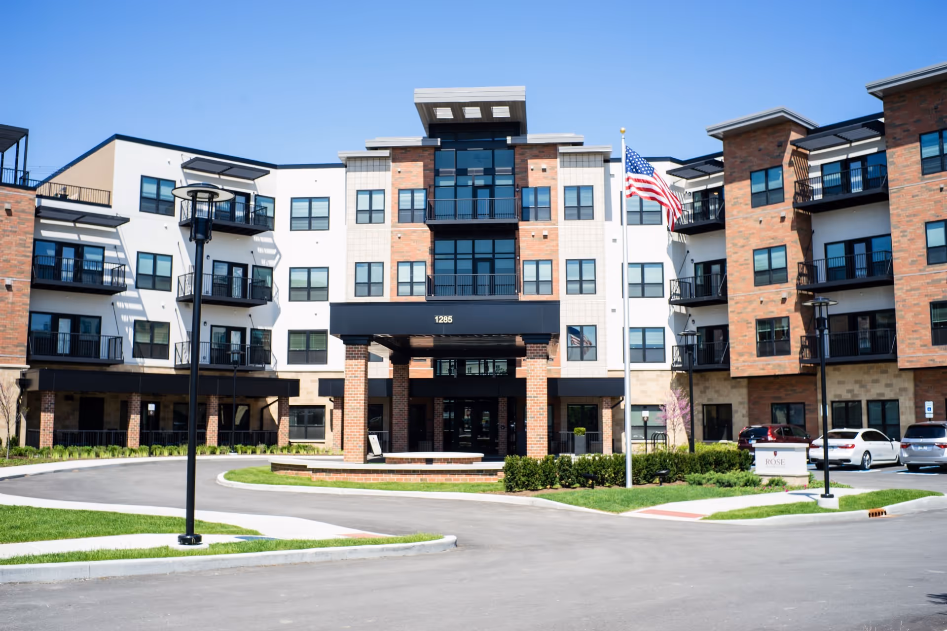 Front exterior view of a modern senior living facility named Rose Senior Living Carmel, featuring a multi-story building with balconies, large windows, a covered entrance, an American flag on a flagpole, and a parking area with several cars.