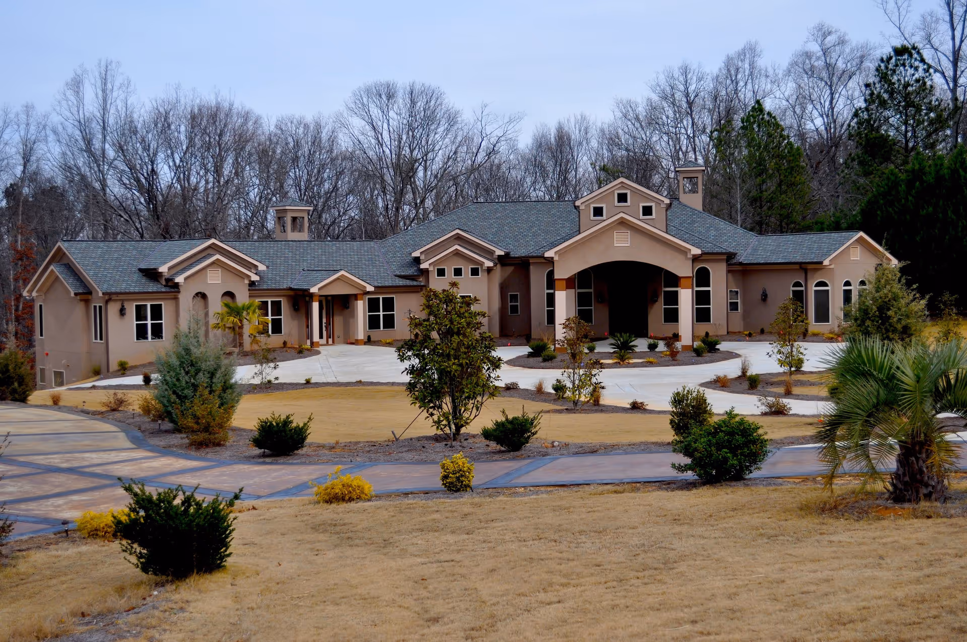 Front exterior view of a single-story senior living facility building with a large driveway and landscaped yard with small trees and bushes. The building has a beige facade with multiple roof peaks and a covered entrance. Leafless trees are visible in the background.