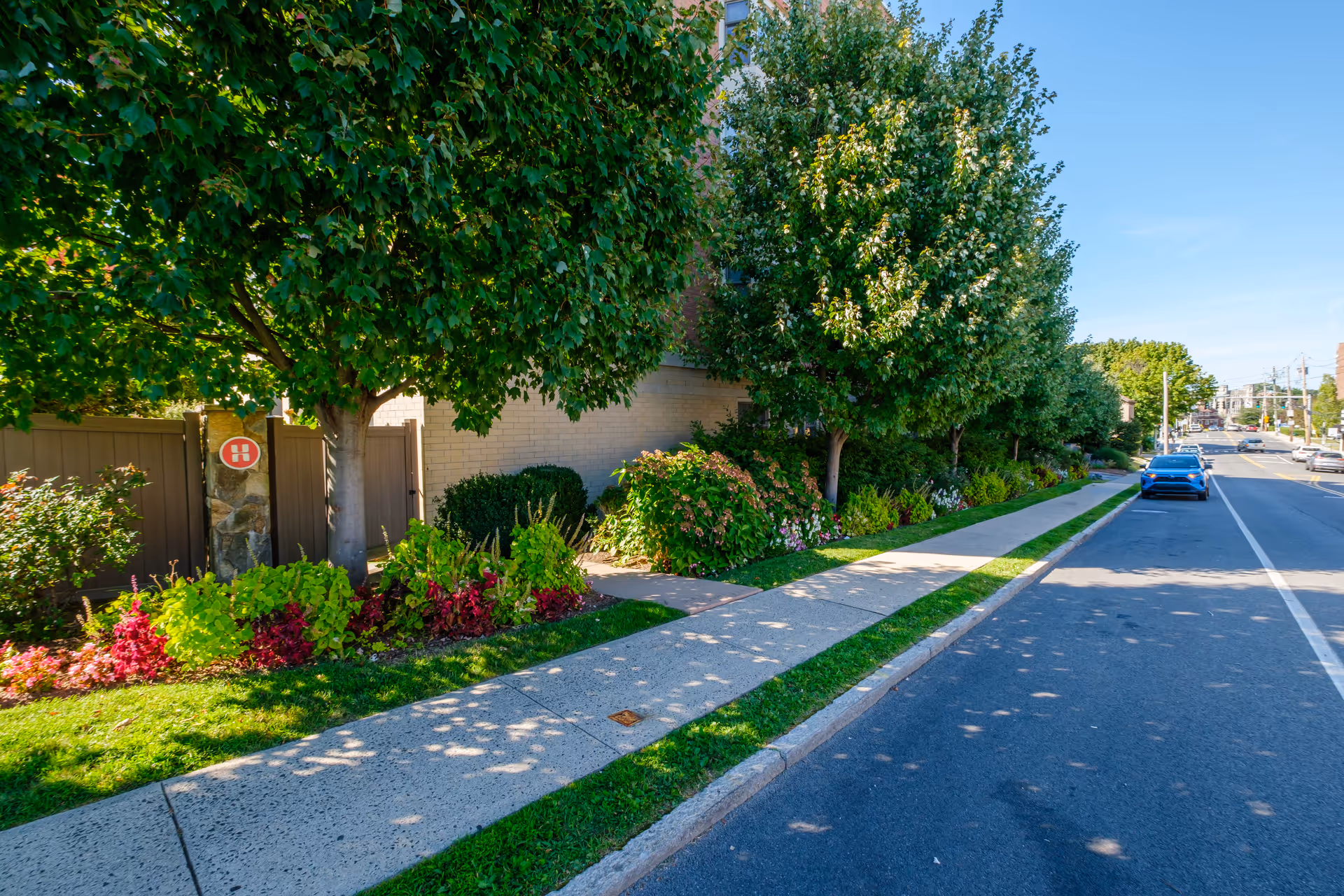 Sidewalk lined with green trees and colorful flower beds next to a brick building under a clear blue sky, with a street and parked cars on the right side.