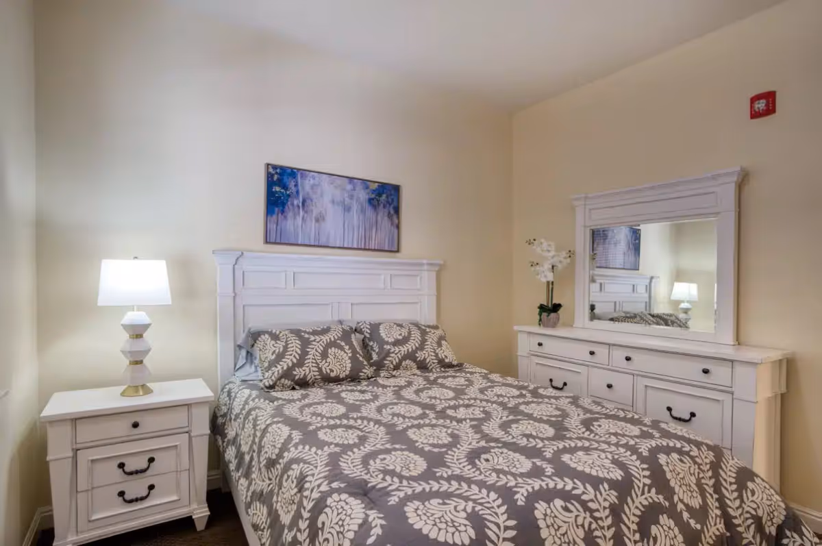 A bedroom with a white wooden bed frame and matching nightstand and dresser. The bed is made with a gray and white patterned comforter and two pillows. A white lamp is on the nightstand, and a vase with white flowers is on the dresser. A framed artwork with blue and gray tones hangs above the bed.