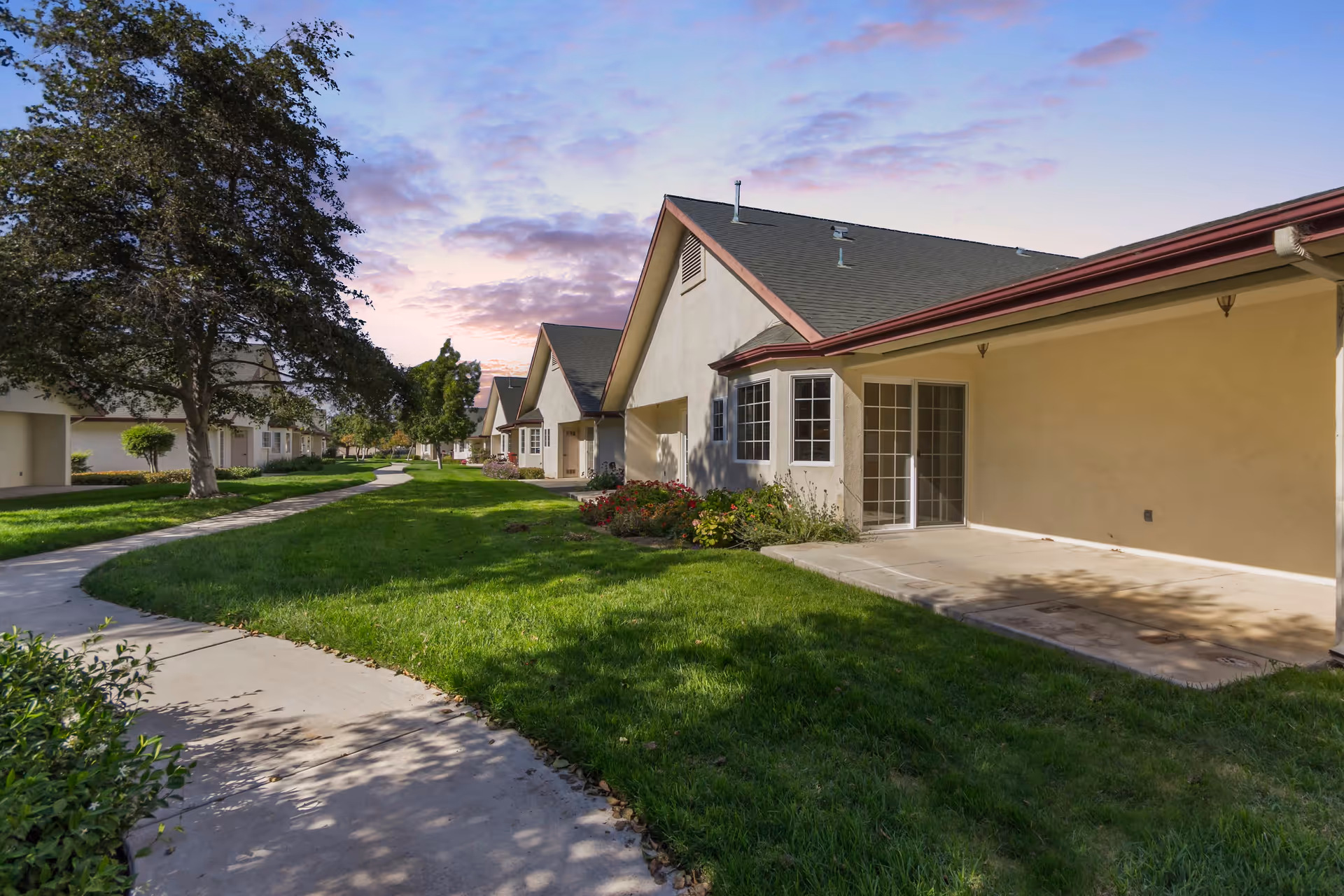 View of a residential senior living community with a paved walkway curving through a grassy area, lined with trees and single-story buildings with beige walls and dark roofs under a partly cloudy sky at sunset.