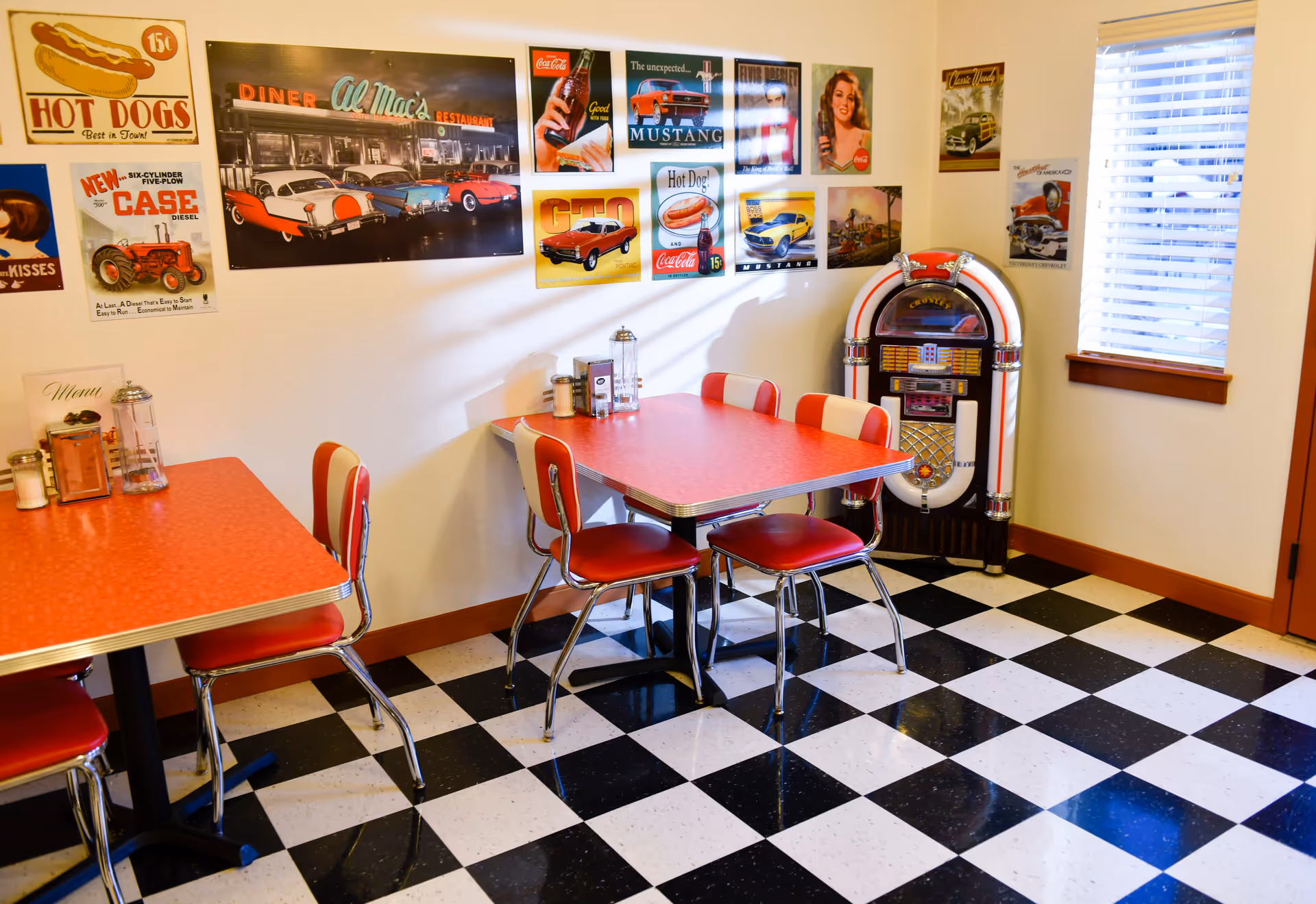 A retro-style dining area with red and white chairs and tables, black and white checkered floor, vintage posters on the walls, and a classic jukebox in the corner near a window with blinds.