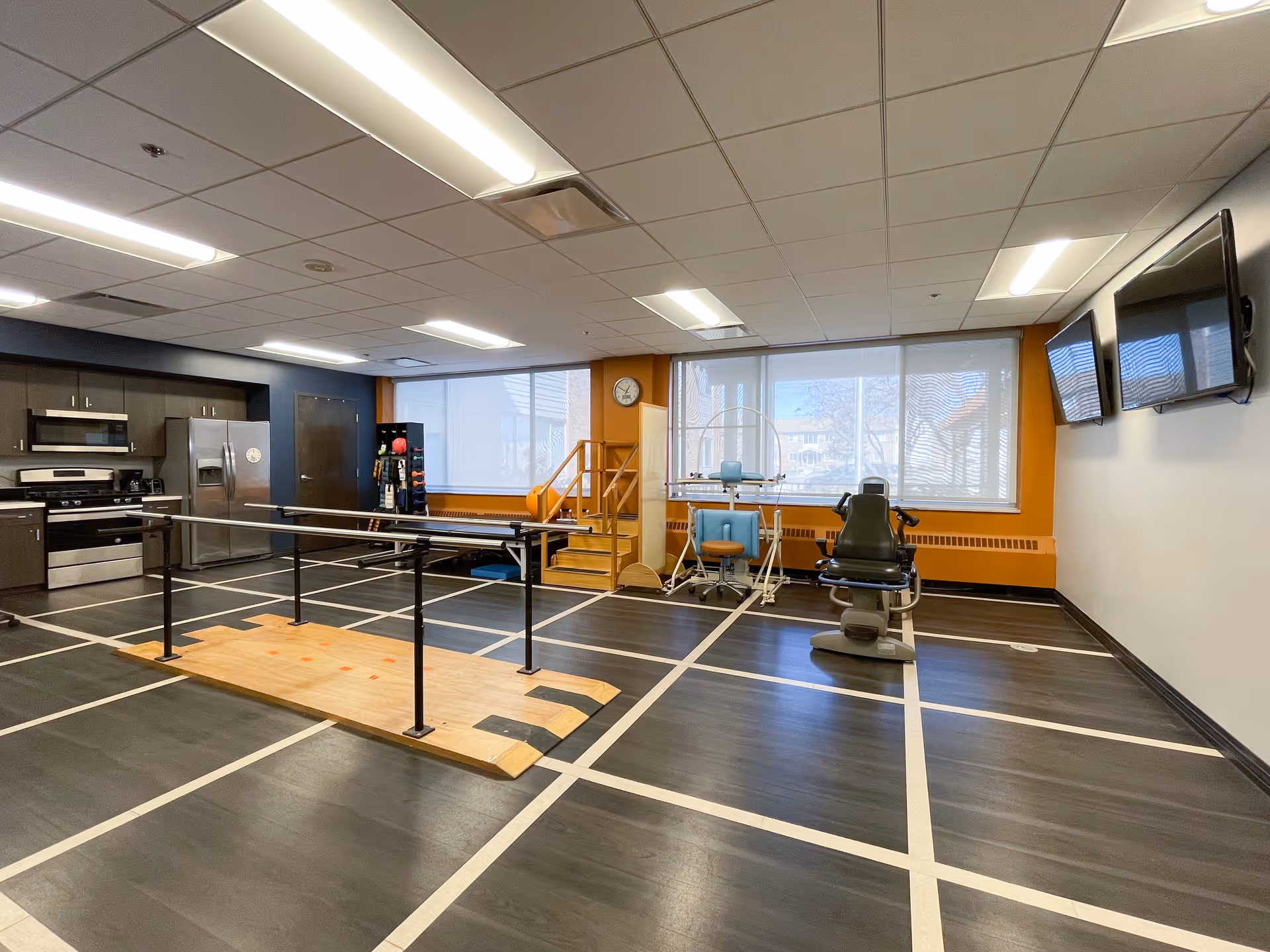 Interior therapy/exercise room with parallel walking bars, rehabilitation equipment, a kitchenette area, and wall-mounted TVs.