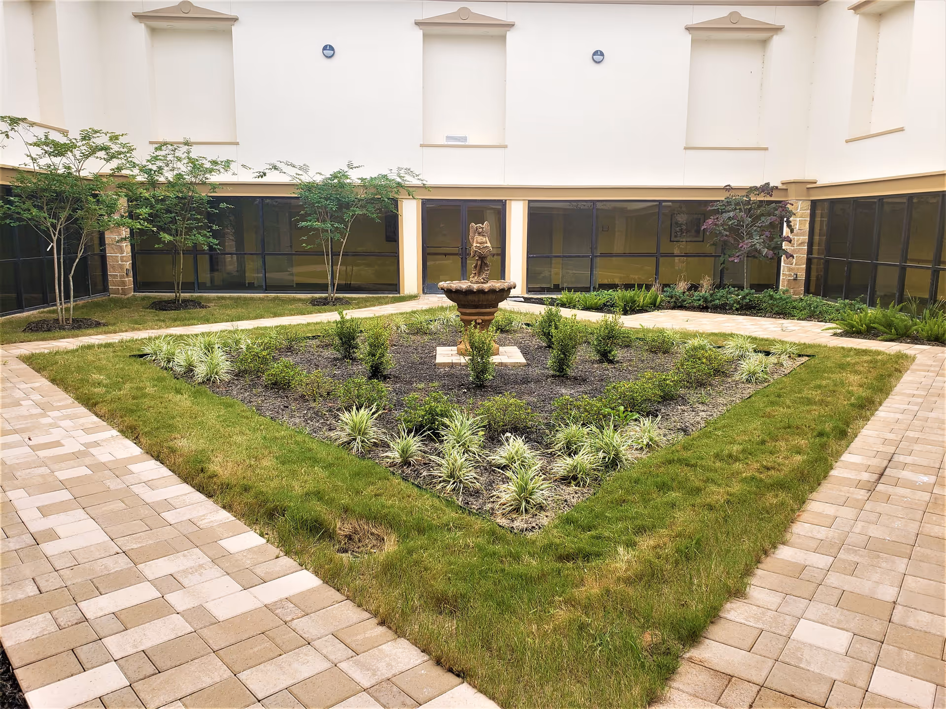 Indoor courtyard garden with a central stone fountain surrounded by neatly trimmed grass, small plants, and trees. The courtyard is enclosed by a building with large windows and beige walls.