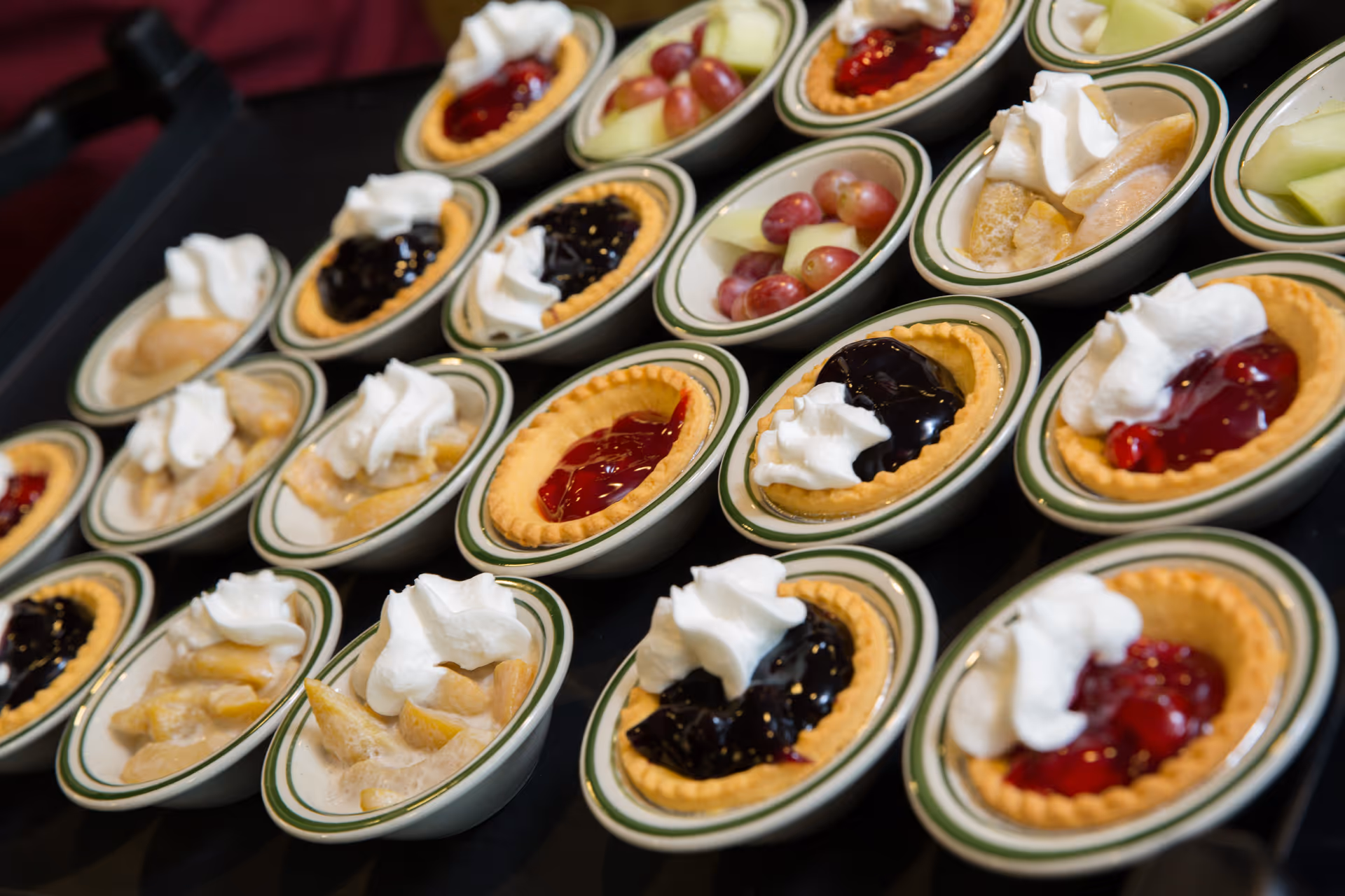Multiple small dessert tarts and bowls of fruit topped with whipped cream arranged on a table.
