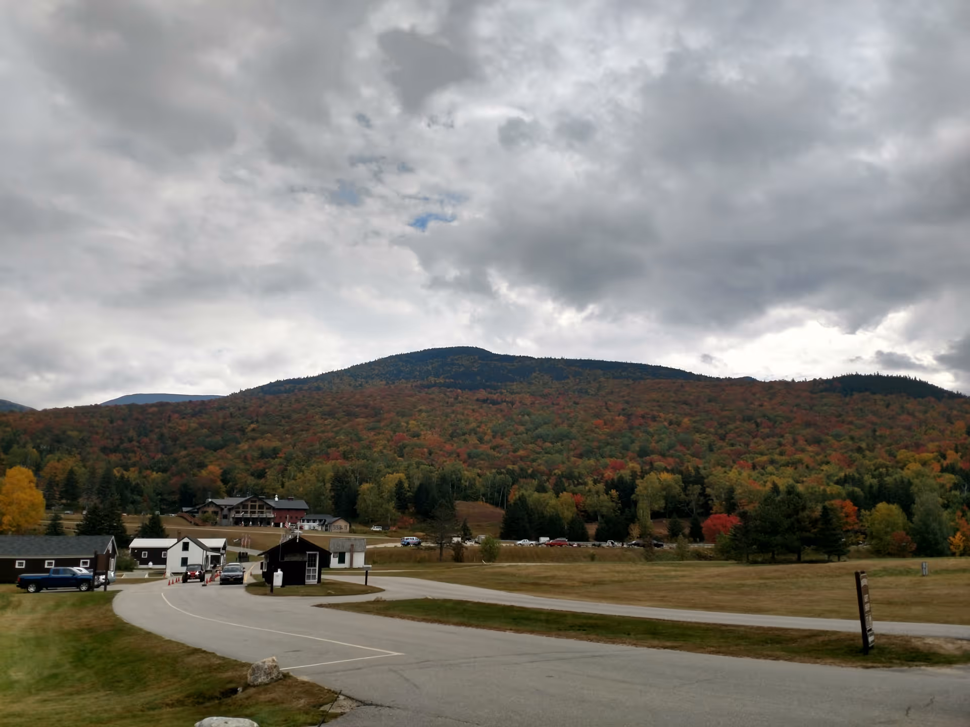 A scenic outdoor view featuring a winding road leading to several small buildings with a backdrop of a forested mountain covered in autumn foliage under a cloudy sky.
