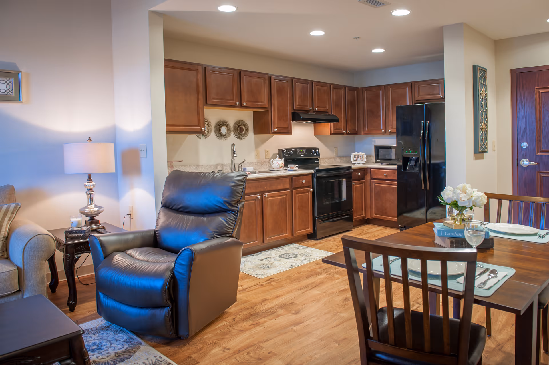 A cozy interior space featuring a kitchen with dark wood cabinets, black appliances including a refrigerator and stove, and a dining area with a wooden table set for two. Adjacent to the kitchen is a living area with a black leather recliner, a beige sofa, a side table with a lamp, and decorative wall art. The flooring is wood, and the space is well-lit with ceiling lights.