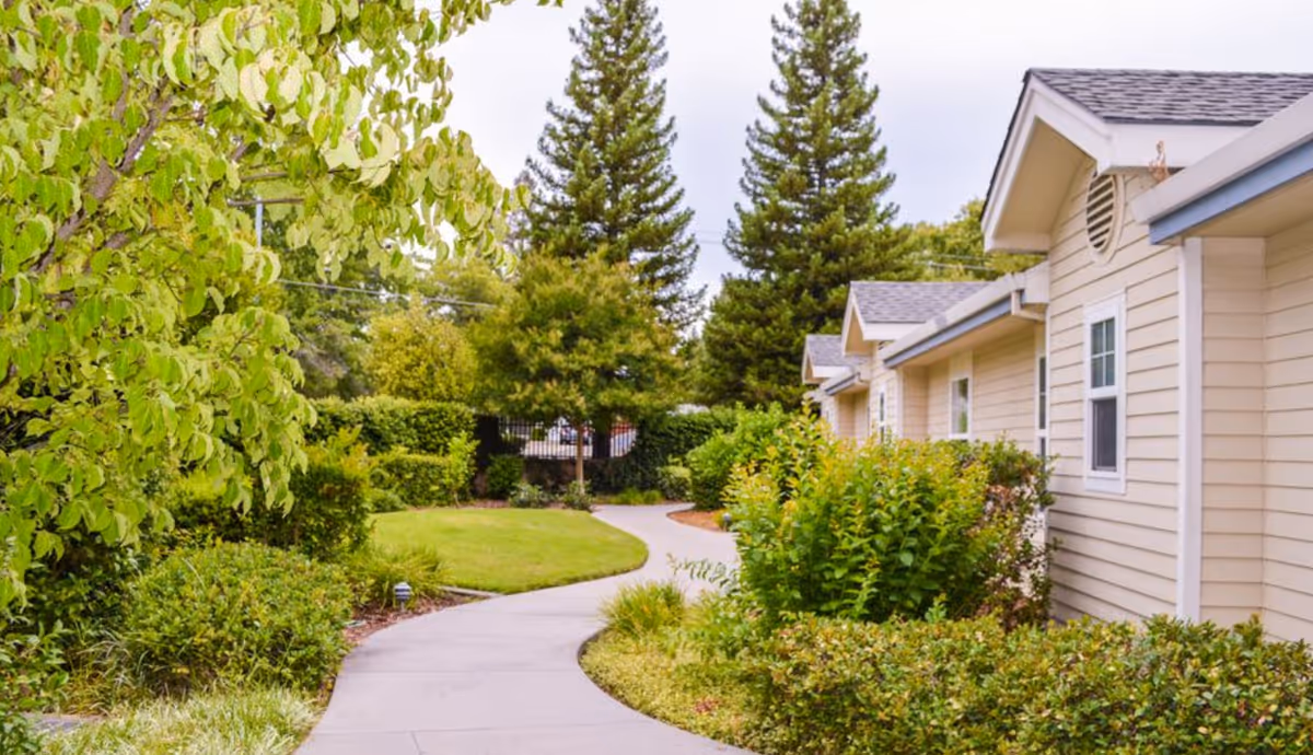 A winding concrete pathway surrounded by green bushes, trees, and grass leading alongside a beige building with white-trimmed windows under a cloudy sky.