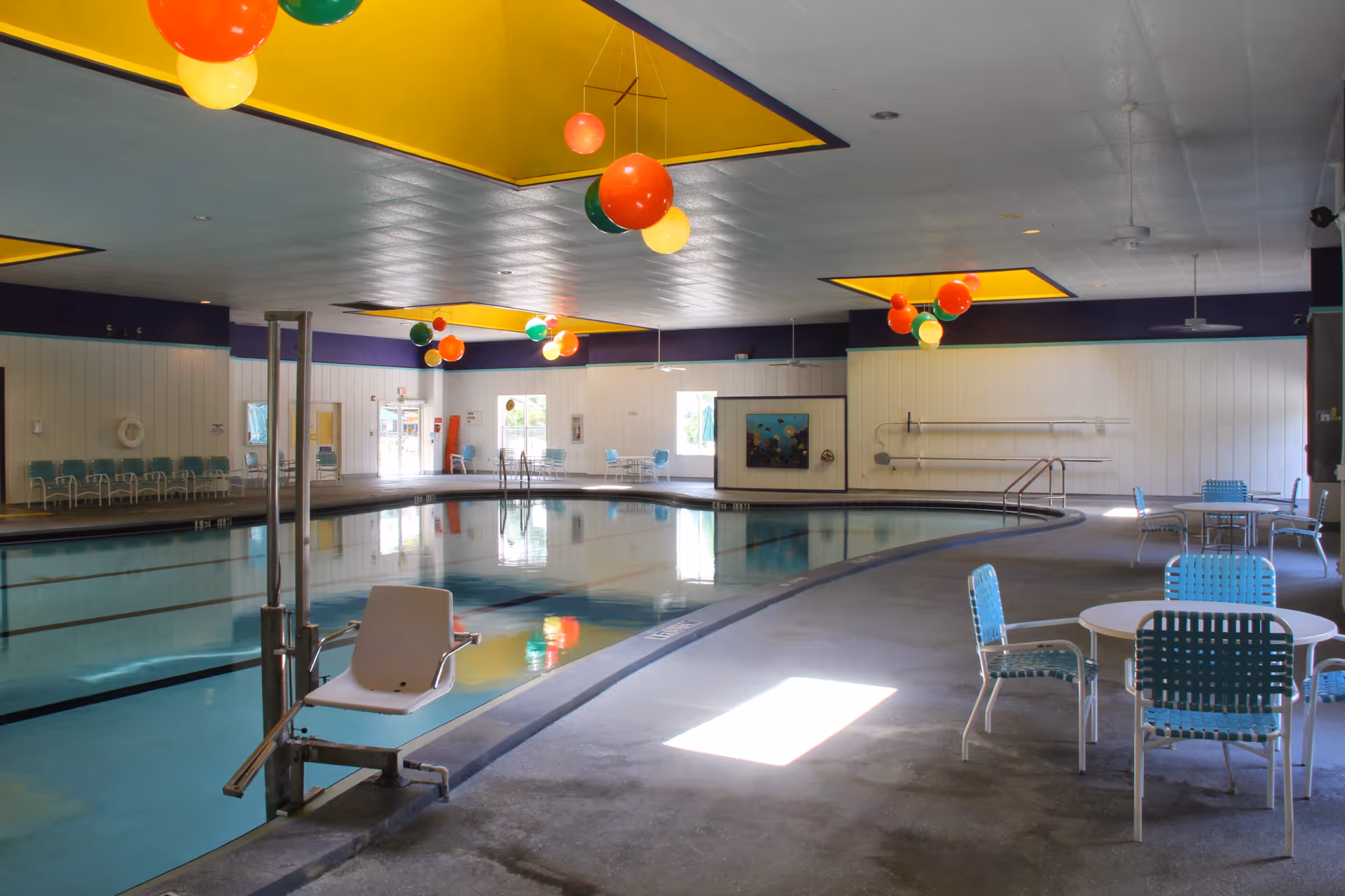 Indoor swimming pool area with tables and chairs, a pool lift, and colorful hanging lights.