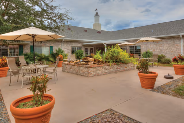 Sunny courtyard patio with umbrellas, tables, chairs, potted plants, and a central raised planter/fountain in front of a brick building.