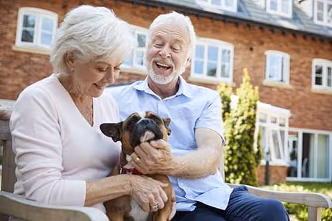 An elderly couple sitting on a bench outside a brick building, smiling and holding a small brown dog between them.