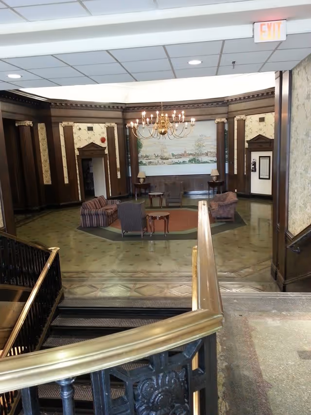 Interior view of a senior living facility lobby area with a staircase in the foreground leading down to a seating area. The seating area includes several upholstered chairs and small tables arranged on a patterned floor. The walls feature dark wood paneling with decorative columns and a large mural painting. A chandelier with multiple lights hangs from the ceiling, and there is an exit sign visible above a doorway.