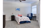 A bright bedroom with a single bed covered with a patterned bedspread and a red pillow. There is a dark wooden nightstand with a lamp and decorative items on the left side of the bed. A blue armchair and a small round table with a lamp are positioned near a window with white blinds. The room has light-colored walls, wood flooring, a ceiling fan, and a framed artwork above the bed.