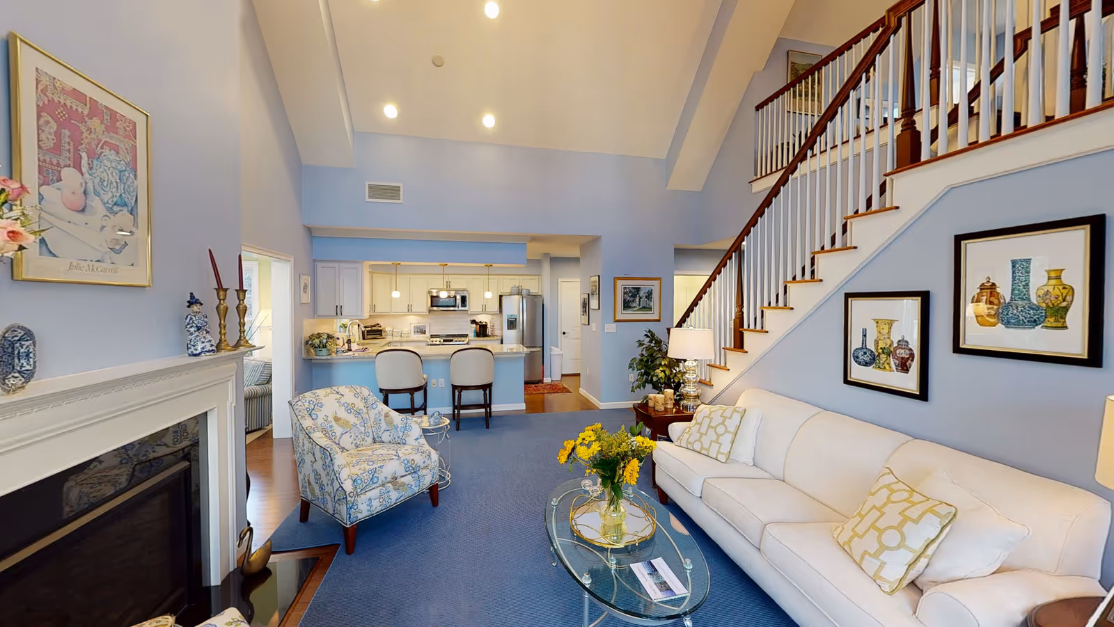 A bright and spacious living room with a white sofa adorned with patterned cushions, a glass coffee table with a vase of yellow flowers, and a floral armchair. The room features a fireplace with decorative items on the mantel, framed artwork on the walls, and a staircase with wooden handrails leading to an upper floor. In the background, there is an open kitchen with a breakfast bar and two chairs.