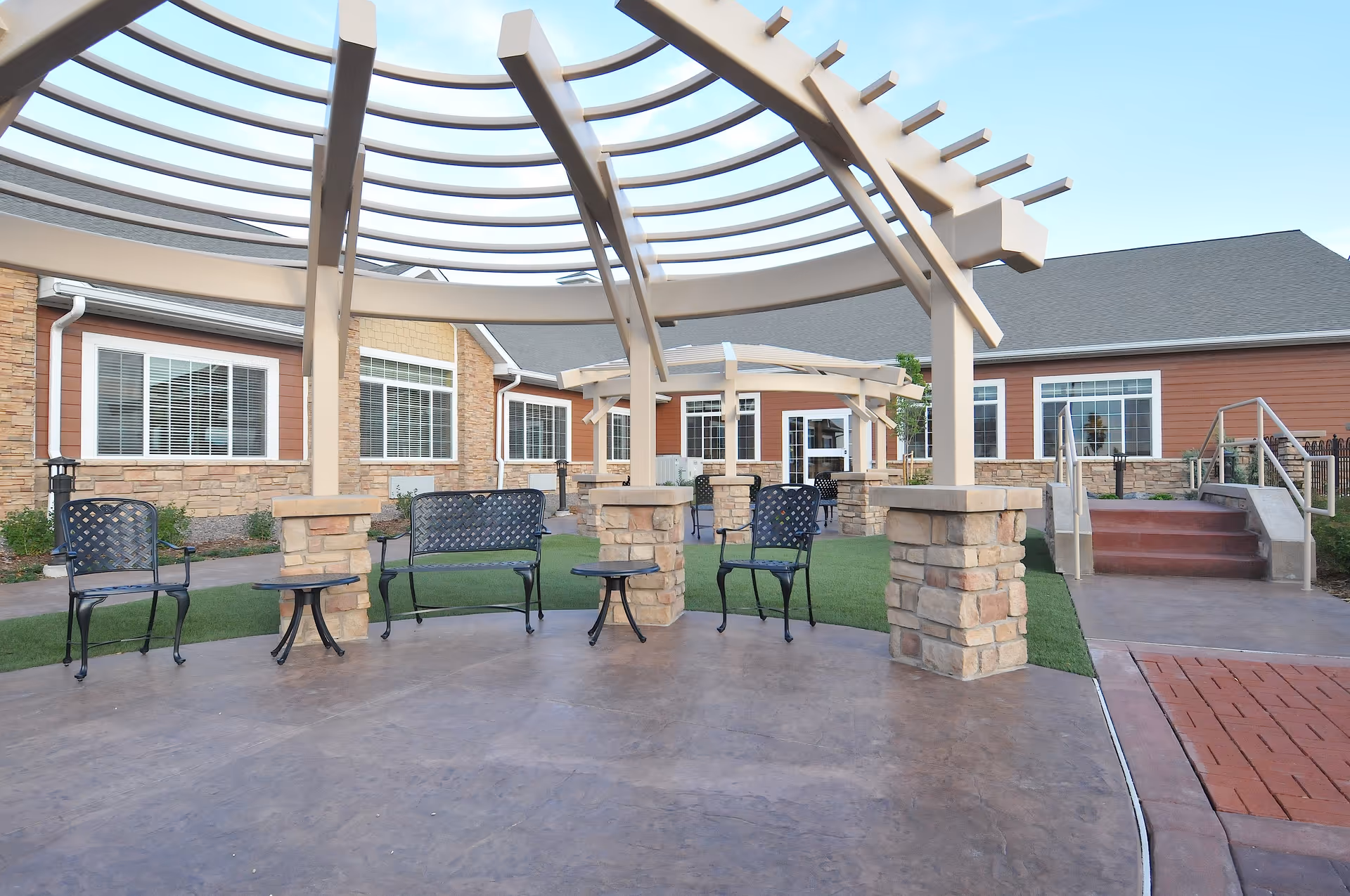 Outdoor seating area at a senior living facility with metal chairs and benches under a large pergola structure. The area is paved with concrete and surrounded by grass and a building with stone and wood siding in the background.